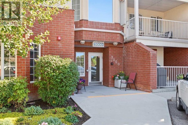 Entrance to apartment building 3000 with red brick and glass double doors. - #, 3308 Sienna Park Green Sw, Calgary