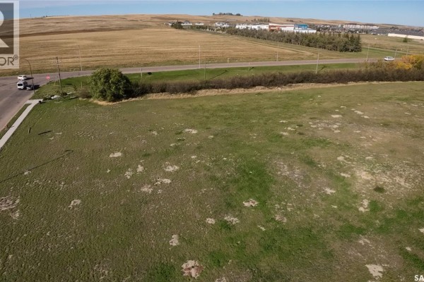 Aerial view of a vacant green field with a road, some trees, and distant buildings. - 01 Springs Drive, Swift Current