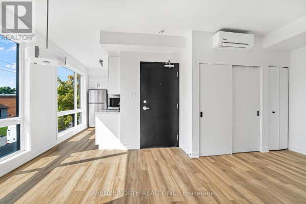 Open concept living space with large windows, light wood floors, and a kitchen area. - 03 - 391 Dieppe Street, Ottawa