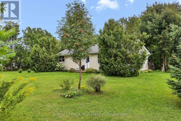 Exterior view of a light-colored bungalow with a dark metal roof, surrounded by green grass and trees. - 046285 Old Mail Road, Meaford