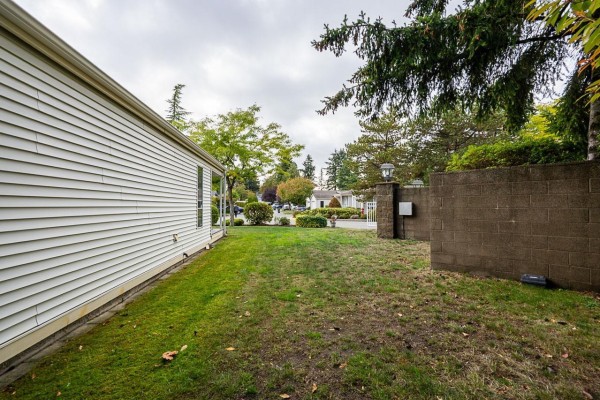 View of a white manufactured home's side exterior and backyard with a stone wall and trees. - 1 2345 Cranley Drive, Surrey