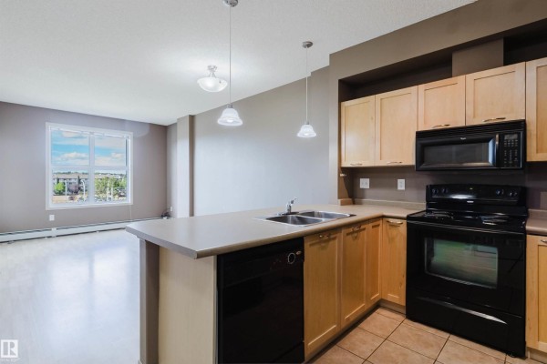 Kitchen with light wood cabinets, beige countertops, black appliances, and adjacent living area. - #1-405 4245 139 Av Nw, Edmonton