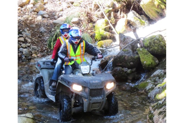 Two people wearing helmets and safety vests ride a green ATV through a shallow, rocky stream. - 1 46891 Sawki Creek Road, Mission