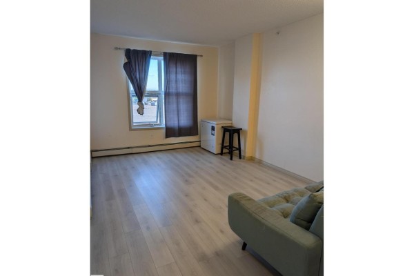 Living area with light wood flooring, dark curtains, and a small freezer. - 1-504, 4245 139 Av Nw Nw, Edmonton
