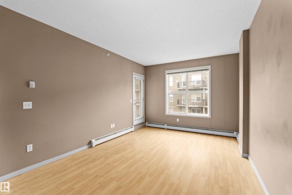 Empty living room with light wood floor, beige walls, and window view of building. - #1-516 4245 139 Av Nw, Edmonton