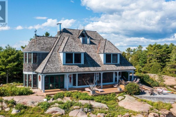 Victorian home with wood shake roof, dormer windows, and wrap-around porch on a rocky waterfront setting.