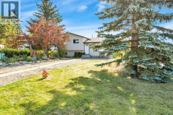 Side view of the beige house with mature evergreen trees and a grassy lawn in front. - 1 Mountain Place, Sylvan Lake