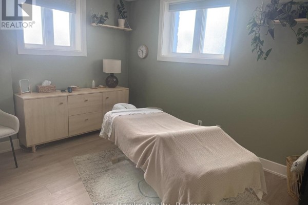 Massage room with a treatment table, light wood cabinet, plants, and two windows. - 1 West - 366 Midland Avenue, Midland