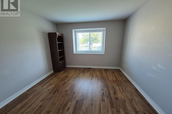 Empty room with light grey walls, wood floor, window, and a dark wood bookshelf. - 10 - 162 First Street, Welland