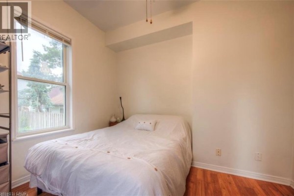 Bedroom with a queen-size bed, white linens, and a large window overlooking the street. - 10 Bruce Street, Brantford
