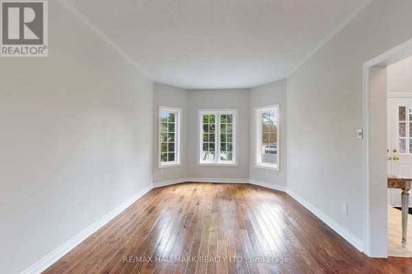 Empty room with large bay windows showcasing lush green trees outside, and dark hardwood floors. - 10 Driftwood Court, Richmond Hill