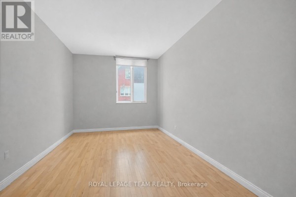 Empty room with light wood flooring, grey walls, and a single window overlooking a brick building. - 10 Lewis Street, Ottawa