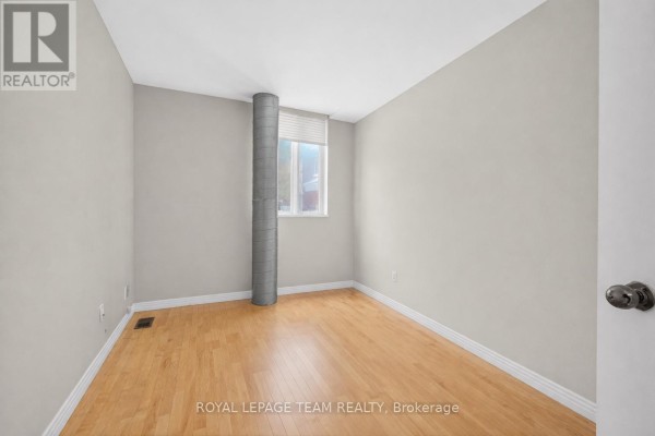 Empty room with light wood flooring, grey walls, a round metal column, and a single window. - 10 Lewis Street, Ottawa