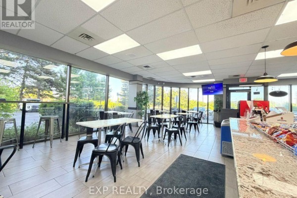 Seating area with tables and chairs beside large windows, with a counter and shelves visible. - #100 - 700 Dorval Drive, Oakville