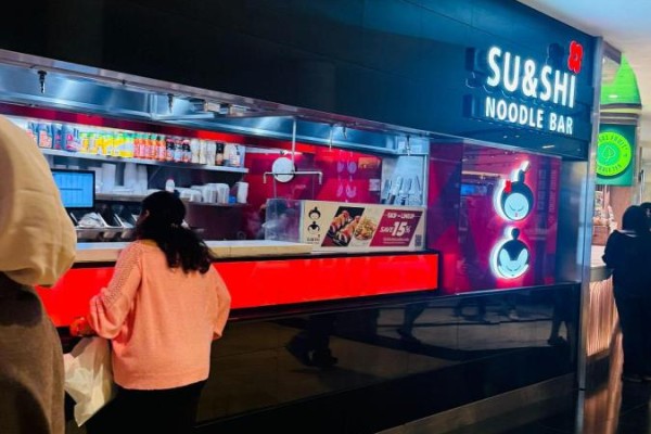 Customers at a sushi and noodle bar counter, ordering food with a view of menu boards. - 100 City Centre Drive, Mississauga