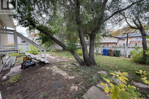 Backyard view with a lawnmower, green and blue bins, and a large tree. - 100 Marshall Crescent, Regina