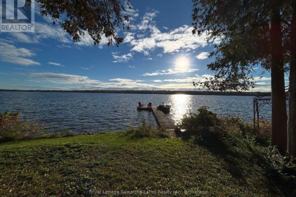 Sunrise over a wide lake with a dock and two red chairs visible from a grassy shore. - 100 Pinewood Boulevard, Kawartha Lakes