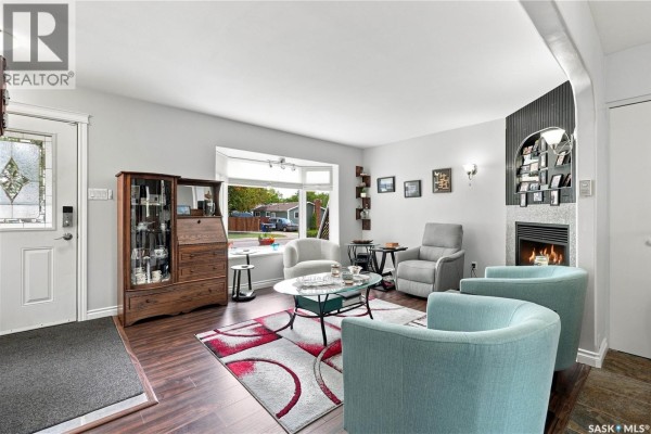Living room with light grey walls, teal armchairs, a bay window, and a gas fireplace with a stone surround. - 1001 Mahoney Avenue, Saskatoon