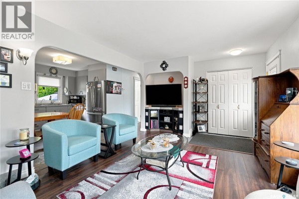 Living room and kitchen area with teal armchairs, a television, and an opening to the kitchen with grey cabinets. - 1001 Mahoney Avenue, Saskatoon