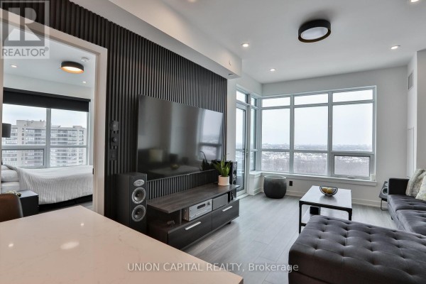 Living room with a large TV, dark sectional sofa, and view of the city through large windows. - 1002 - 30 Upper Mall Way, Vaughan