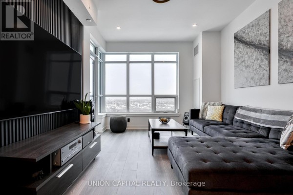 Living room featuring a dark grey sectional sofa, coffee table, and expansive city view. - 1002 - 30 Upper Mall Way, Vaughan