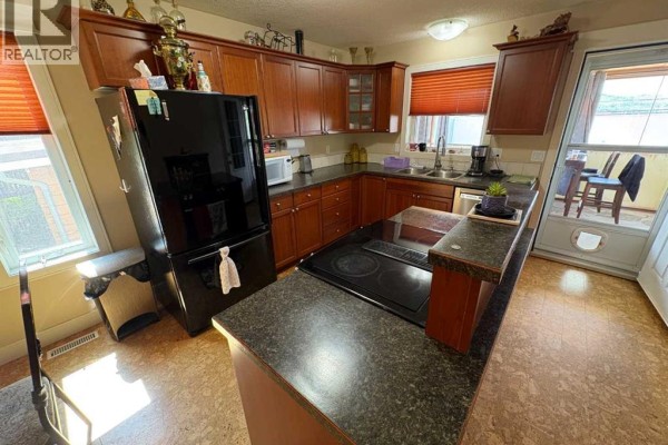 Kitchen features dark wood cabinets, black refrigerator, stainless steel dishwasher, and a door leading to a patio. - 10032 102 Street, Peace River