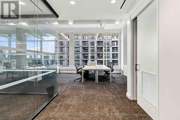 Hallway view of an office with glass-walled offices, desks, chairs, and carpeted flooring. - 1005 - 130 Queens Quay E, Toronto
