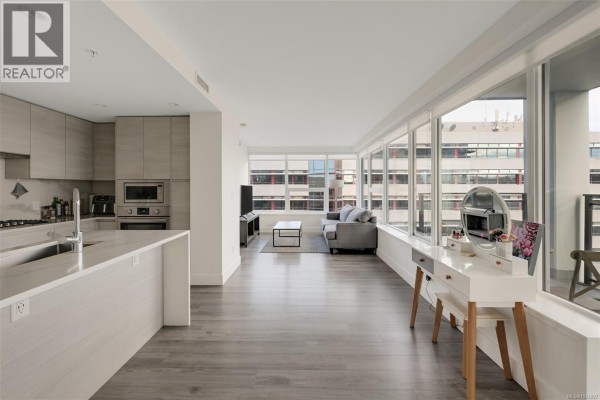 Open concept living area and kitchen with large windows, grey sofa, and vanity table. - 1006 777 Herald St, Victoria