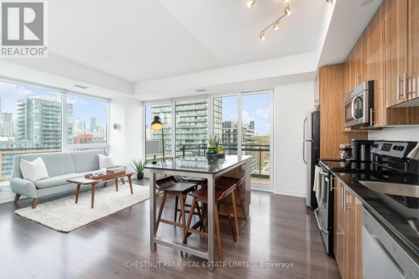 Open concept living and kitchen area with island, stainless steel appliances, and wooden cabinets. - 1009 - 225 Sackville Street, Toronto