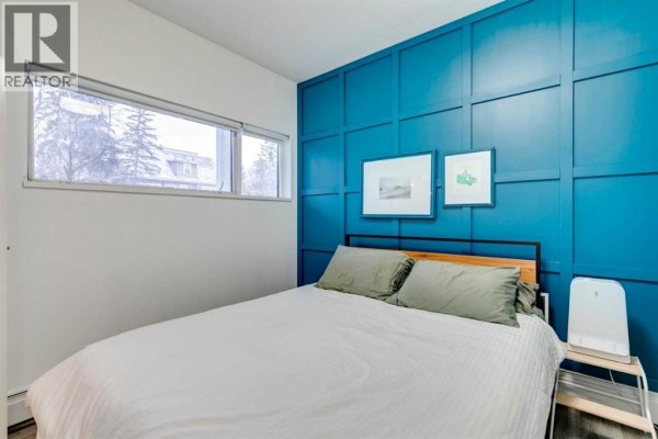 Bedroom with a bed, window overlooking trees, blue paneled accent wall, and light wood flooring. - 101, 1521 26 Avenue Sw, Calgary