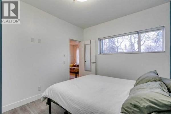 Bedroom with a bed, white walls, window overlooking trees, and an open doorway. - 101, 1521 26 Avenue Sw, Calgary