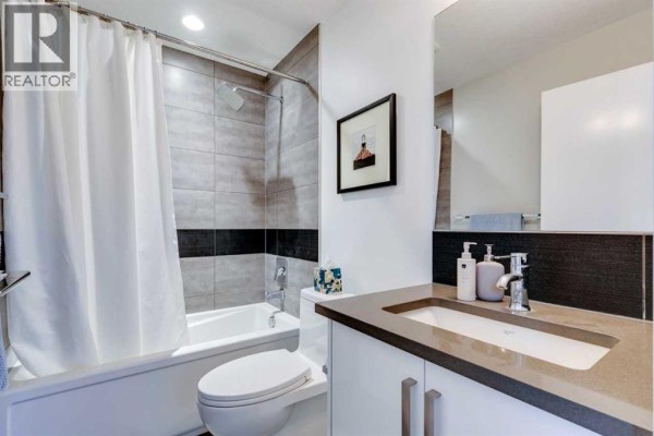 Bathroom with a white bathtub, shower curtain, vanity, and gray tile accents. - 101, 1521 26 Avenue Sw, Calgary