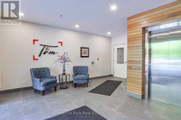 Apartment building lobby with grey tiled floor, two blue upholstered chairs, and a stainless steel elevator. - 101 - 23 Campus Trail, Huntsville
