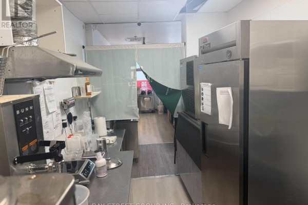 Commercial kitchen interior with stainless steel counters, appliances, refrigerator, and a green fabric partition. - 101 - 330 Highway 7 E, Richmond Hill