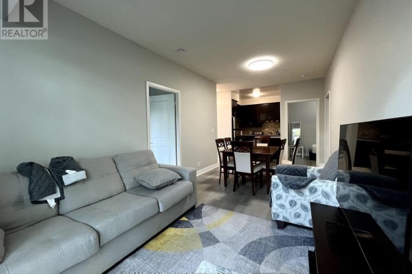 Living room with grey sofa, patterned armchair, TV, and dining area beyond. - 101 - 80 Absolute Avenue, Mississauga