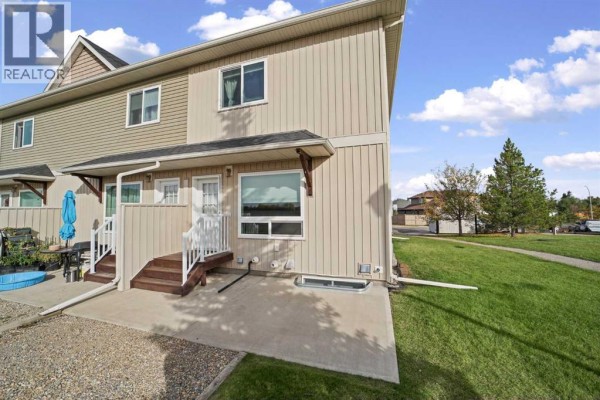 Back of a townhouse with a concrete patio, green lawn, and wooded area. - 101 Clover Way, Carstairs