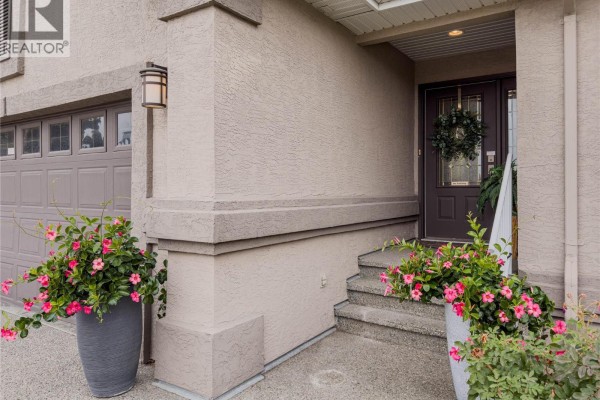 Front entrance with a dark brown door, side windows, and planters with pink flowers. - 101 Coppertree Court, Kamloops