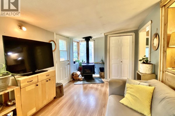 Living room with a wood-burning stove, large television, light wood cabinets, and a grey sofa with yellow cushions. - 1010 Main Street, Valemount