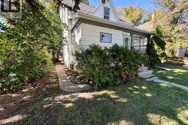 Side exterior view of the house with a pathway and dense green shrubbery. - 1014 6th Avenue Nw, Moose Jaw