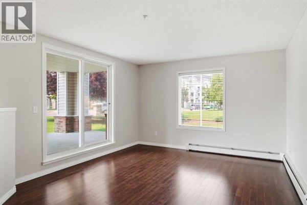 View of empty living room with light gray walls, dark hardwood floors, and large windows. - 102, 17 Country Village Bay Ne, Calgary