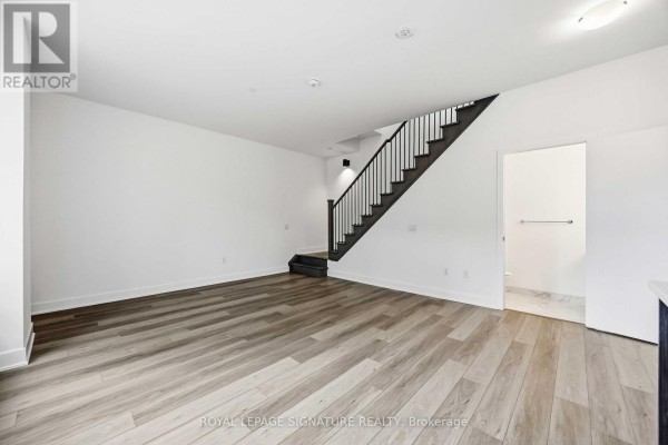 Empty living space with white walls, dark wood staircase, and light wood-look flooring. - 102 - 2 Teagarden Court N, Toronto