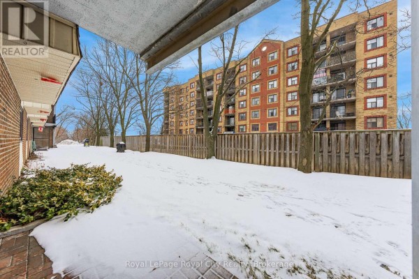 Ground-level view of the backyard with snow, fence, and adjacent building - 102 - 234 Willow Road, Guelph