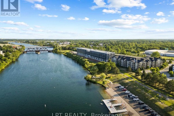 Aerial view of an apartment building complex by a river with a parking lot and green space. - 102 - 350 Prince Charles Drive S, Welland