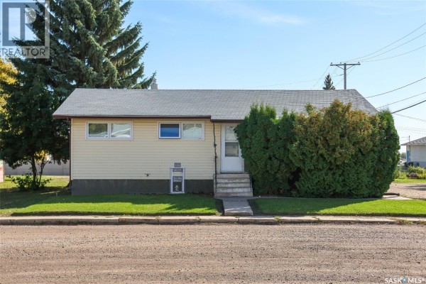 View of the pale yellow bungalow's front entrance with concrete steps and manicured bushes. - 102 4th Avenue E, Blaine Lake