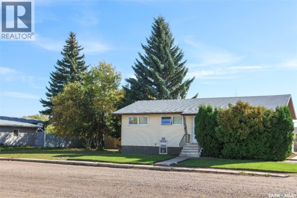The bungalow's front exterior featuring large evergreen trees and a light yellow siding. - 102 4th Avenue E, Blaine Lake