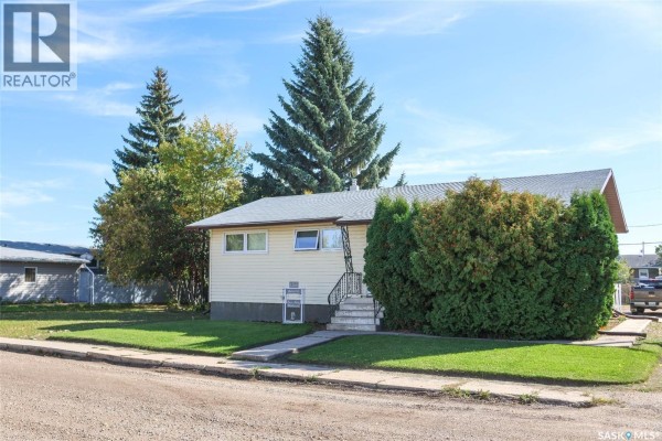 A residential bungalow with light yellow siding, a grey roof, and mature trees in the yard. - 102 4th Avenue E, Blaine Lake