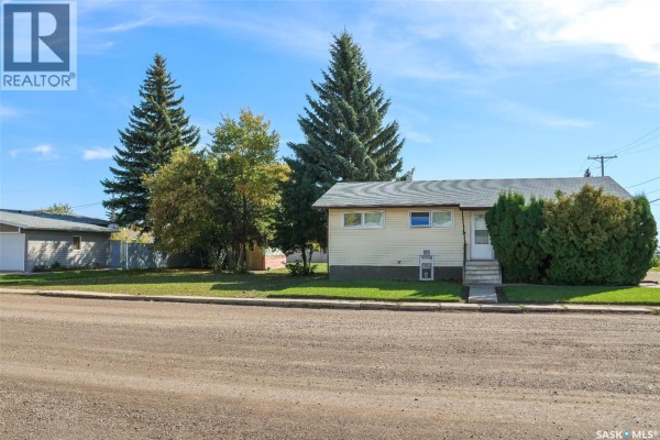 Exterior of a pale yellow home with a grey roof, surrounded by green grass and trees. - 102 4th Avenue E, Blaine Lake