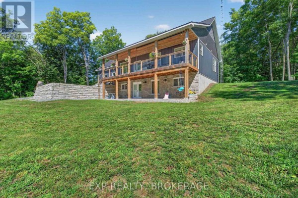 Rear view of the house showing a walk-out basement with stone foundation and a wooden deck above, overlooking a grassy lawn. - 1020 Hill Road, Frontenac