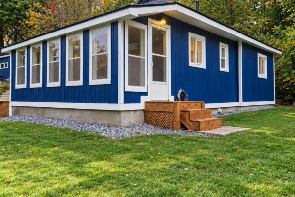 Side view of a blue cottage with multiple windows, a wooden deck, and white trim on a gravel foundation. - 1020 Kellar Road, Frontenac
