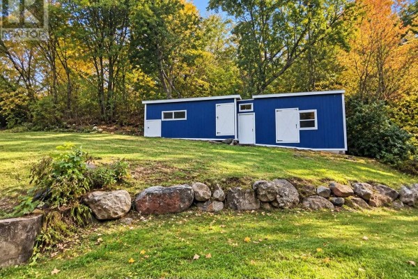 Wide shot of the blue structure with white doors and windows on a sloped lawn with large rocks. - 1020 Kellar Road, Frontenac